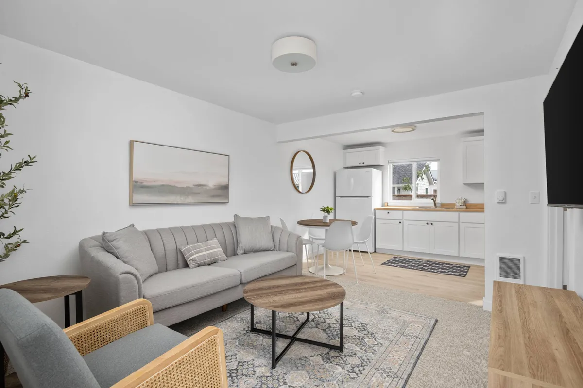 The image showcases a beautifully remodeled condo interior with a plush sofa and round coffee table set in a sunlit living area. The inviting space includes a modern kitchenette, stylish mirror, and tasteful artwork, all creating a cozy, intimate environment ideal for couples. Soft morning light streams in, highlighting the warmth and comfort of the room.