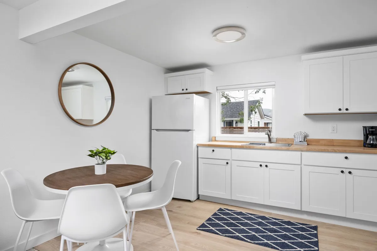 The image displays a round wooden dining table surrounded by white chairs, adjacent to a kitchen countertop and a window with a scenic view. The setup is airy and cozy, ideal for casual dining, coffee breaks, or enjoying a morning routine. The arrangement suggests a seamless transition between meal prep and a comfortable dining experience.