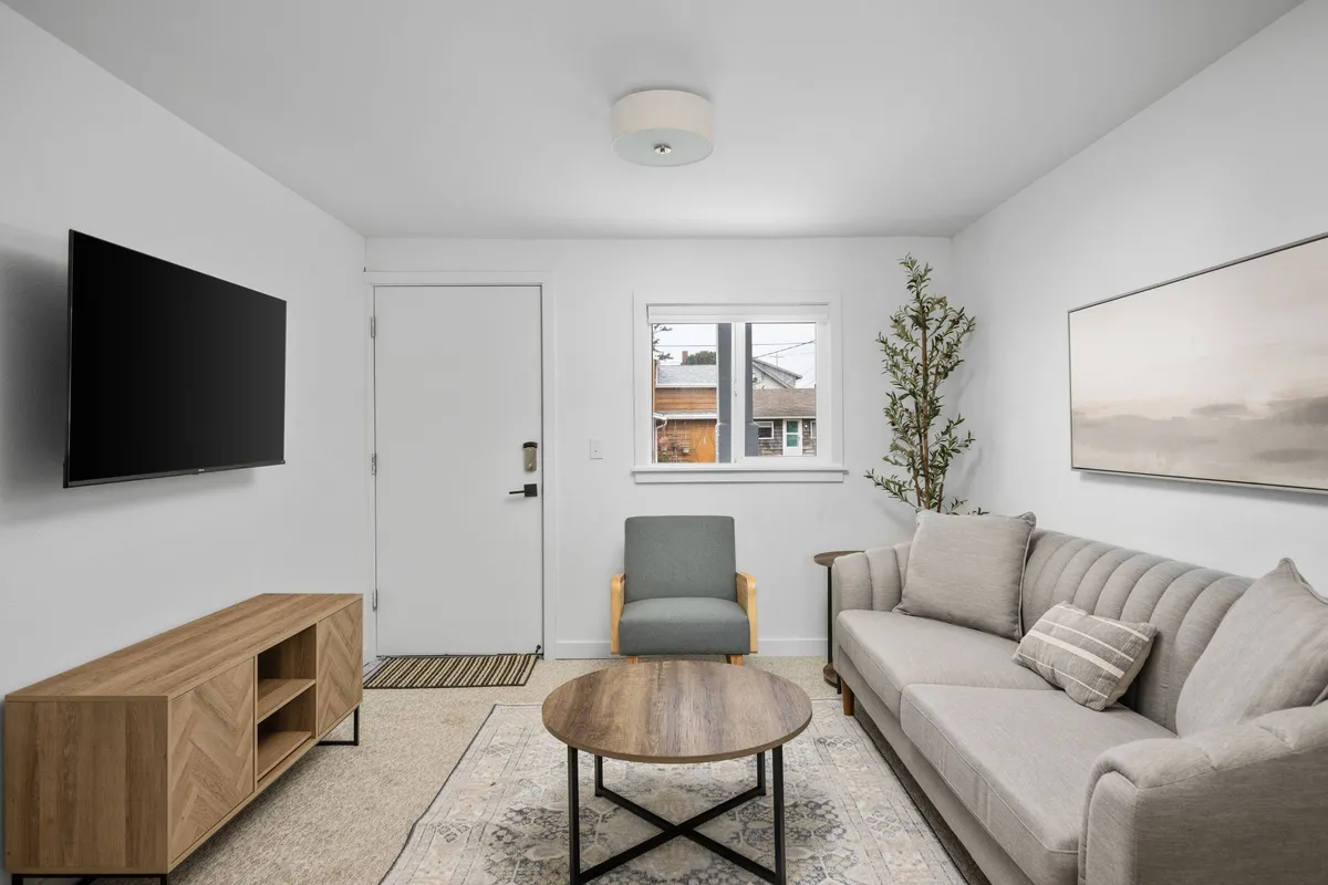 A cozy living area featuring a comfortable sofa, a coffee table, a television, and a large window with a potted plant nearby. The space is arranged to feel both inviting and functional, making it easy for guests and their pets to relax or head outside for a walk.