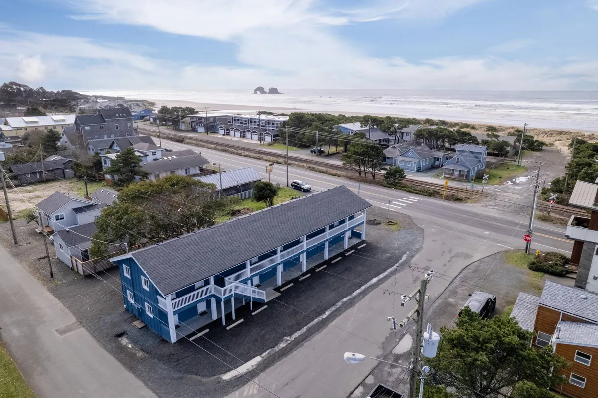 This image showcases the iconic blue building of the Inn at Rockaway Beach set against an expansive ocean view. The surrounding houses and community elements establish a sense of place, while the beach is visible just steps away, making it clear how close guests are to the shoreline. The sky hints at the possibility of a beautiful sunset, inviting romantic strolls and relaxation.