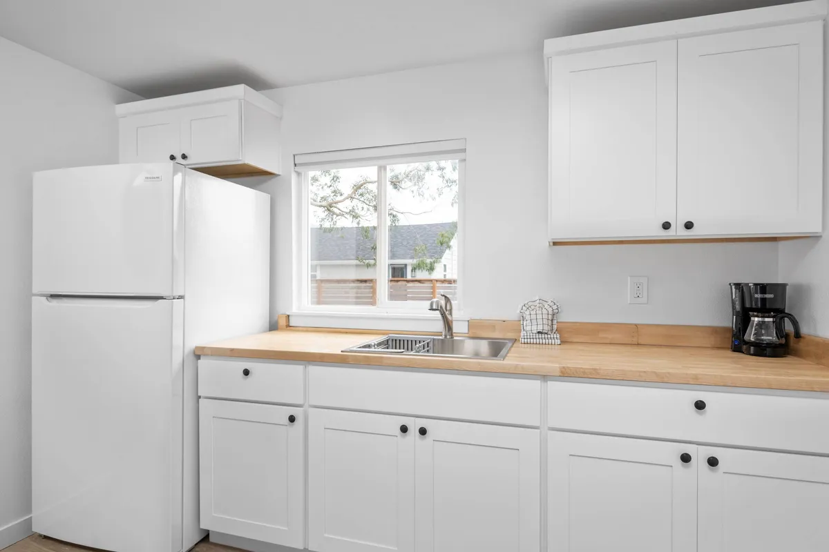 This image features a welcoming kitchenette with a white refrigerator, kitchen sink, and a wood countertop positioned beneath a window that brings in natural light. The scene is inviting and functional, showcasing the core elements needed for light meal prep. The visible appliances and open layout suggest an ideal space for making coffee, preparing breakfast, or a quick snack.