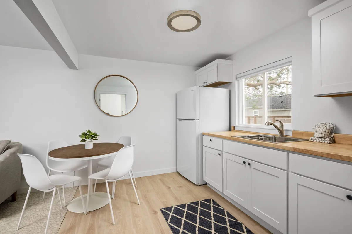This image offers another view of a modern, cozy kitchenette, featuring a round dining table with white chairs, a kitchen sink, fridge, and a mirror, all set in a bright environment. The space is functional yet intimate, encouraging both meal prep and relaxed social interactions.