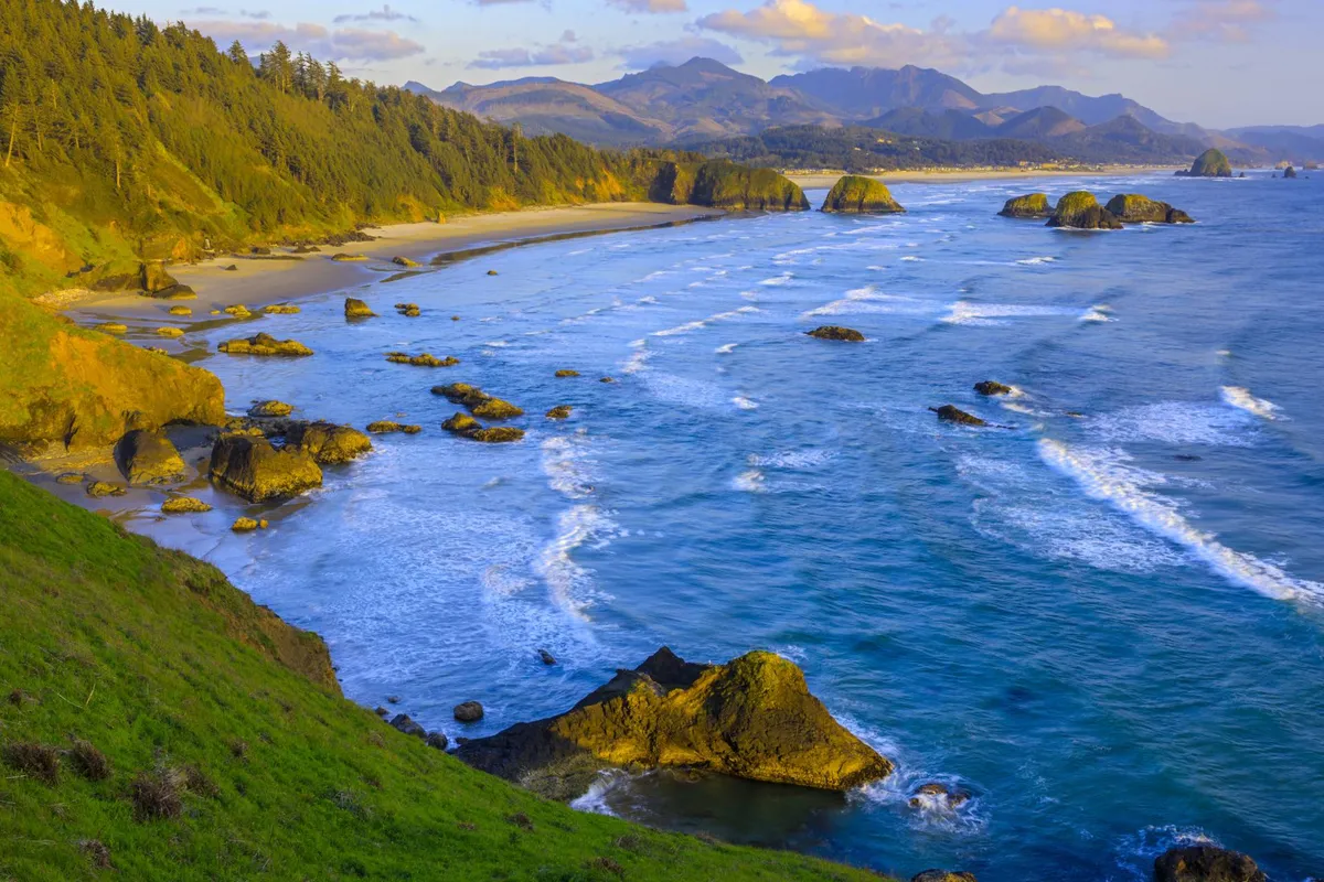 A sweeping view of Rockaway Beach’s rocky coastline, gentle ocean waves, and distant mountains. The lush green hills and wide horizon highlight the natural beauty and serenity of the area, while the soft evening light suggests a perfect setting for sunset strolls, beach walks, and wildlife watching.